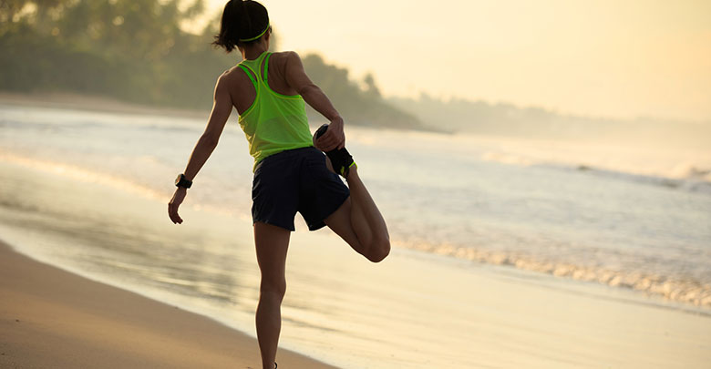 The runner on the beach is stretching her quadriceps to avoid knee injury